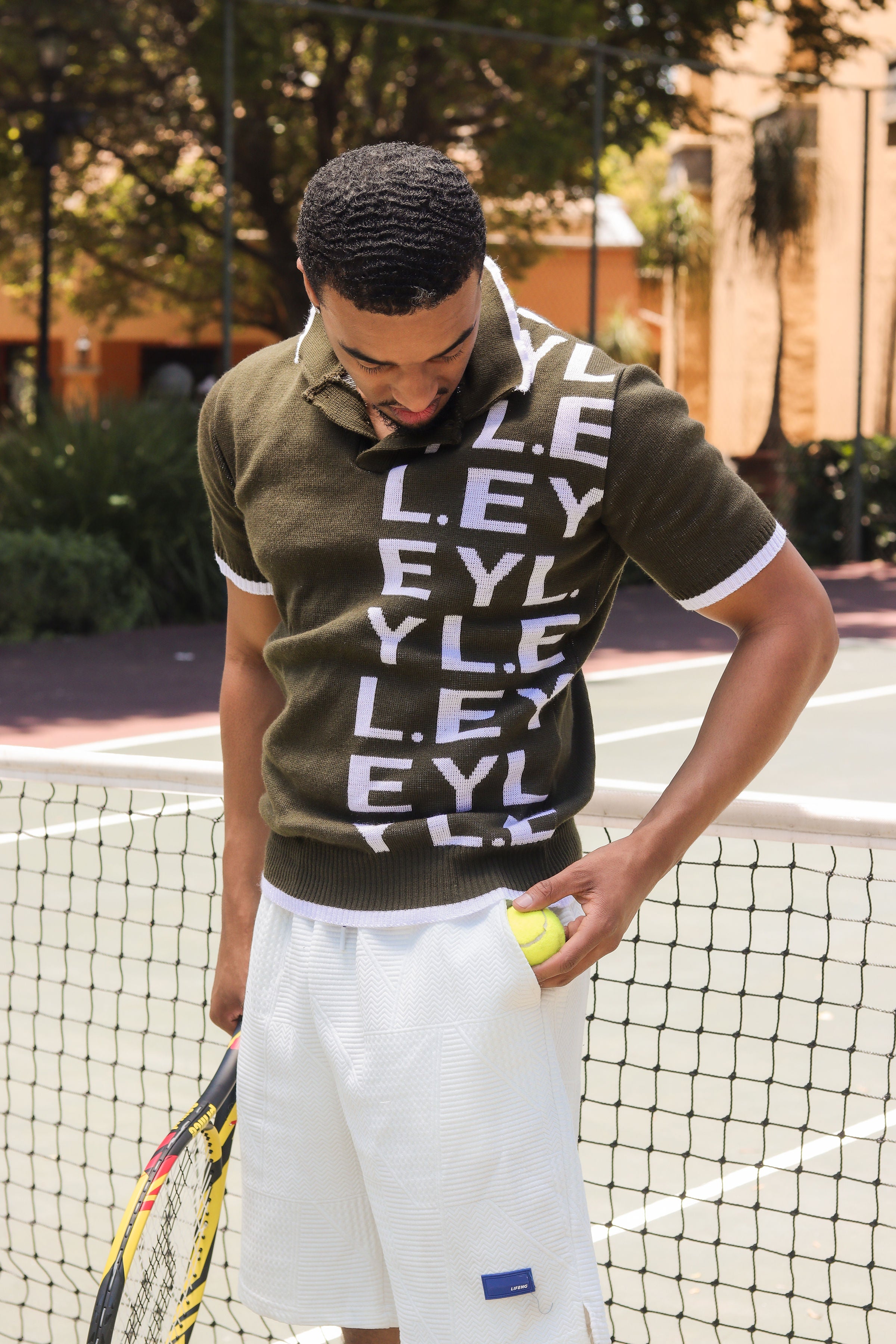 Man on a tennis court wearing a green polo shirt with 'EYL' pattern and white shorts, holding a tennis ball and racket.